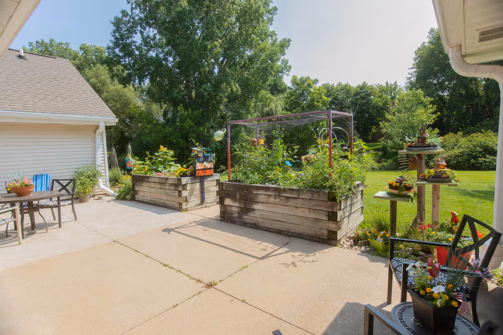 Outdoor patio area with raised garden beds filled with plants and flowers, a small table with chairs, and a multi-tiered plant stand. The background shows green trees and a well-maintained lawn under a clear sky.