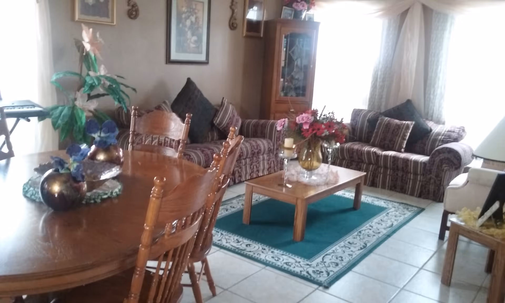 A cozy living room with two patterned sofas adorned with multiple cushions, a wooden coffee table with a vase of pink and red flowers, and a green area rug underneath. To the left, there is a wooden dining table with chairs and decorative vases with blue flowers. The room has tiled flooring, light-colored curtains, framed artwork on the walls, and a wooden cabinet with glass doors in the corner.