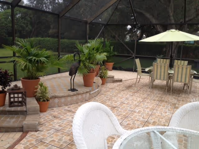 A screened outdoor patio area with tiled flooring, several potted plants, a metal bird sculpture, and outdoor furniture including a glass-top table with white wicker chairs and a dining table with striped chairs under a green umbrella.