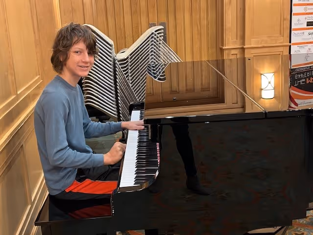 A young person with medium-length brown hair wearing a blue long-sleeve shirt and black and red pants is sitting at a black grand piano in a wood-paneled room with stacked chairs in the background.