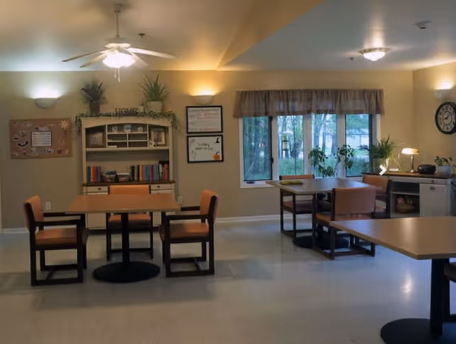 A well-lit common area in a senior living facility featuring several tables with chairs arranged around them. The room has beige walls with wall-mounted lights, a ceiling fan with a light, and large windows with a view of trees outside. There is a cabinet with books and decorative plants, a bulletin board, and a clock on the wall.