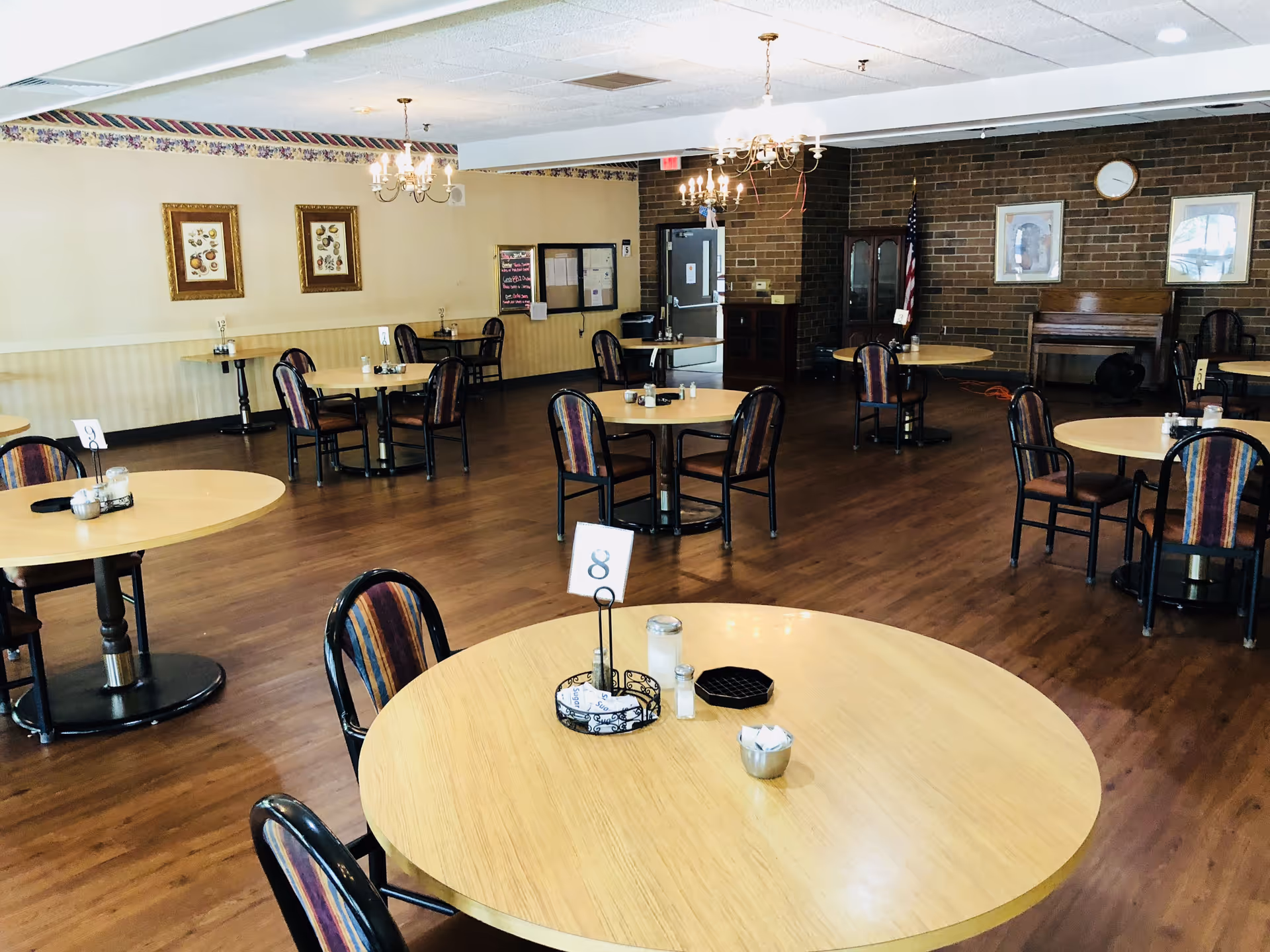 Spacious dining room with round wooden tables and chairs arranged on hardwood floors and chandeliers overhead.