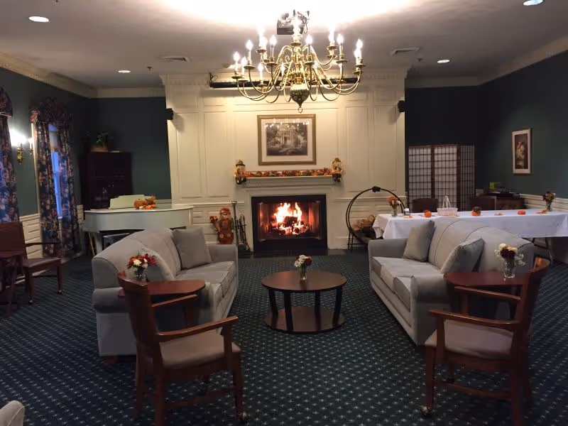 A cozy living room area with two gray sofas facing each other, a round wooden coffee table in the center, and two wooden armchairs. A fireplace with a burning fire is set in a white paneled wall, decorated with autumn-themed ornaments. A chandelier hangs from the ceiling, and there is a white piano in the corner near windows with floral curtains. A long table covered with a white tablecloth and small pumpkins is visible in the background.