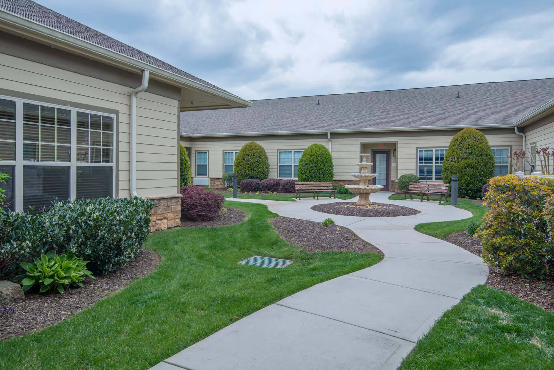 Outdoor courtyard area at Morning Pointe of Hixson featuring a concrete walkway, green grass, landscaped bushes, a three-tiered stone fountain in the center, and benches along the building walls under a cloudy sky.