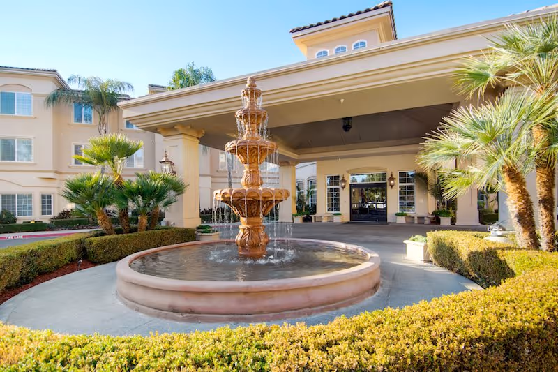 Outdoor view of the entrance to a senior living facility with a large tiered water fountain in the foreground, surrounded by neatly trimmed bushes and palm trees. The building has a covered entryway with columns and multiple windows.