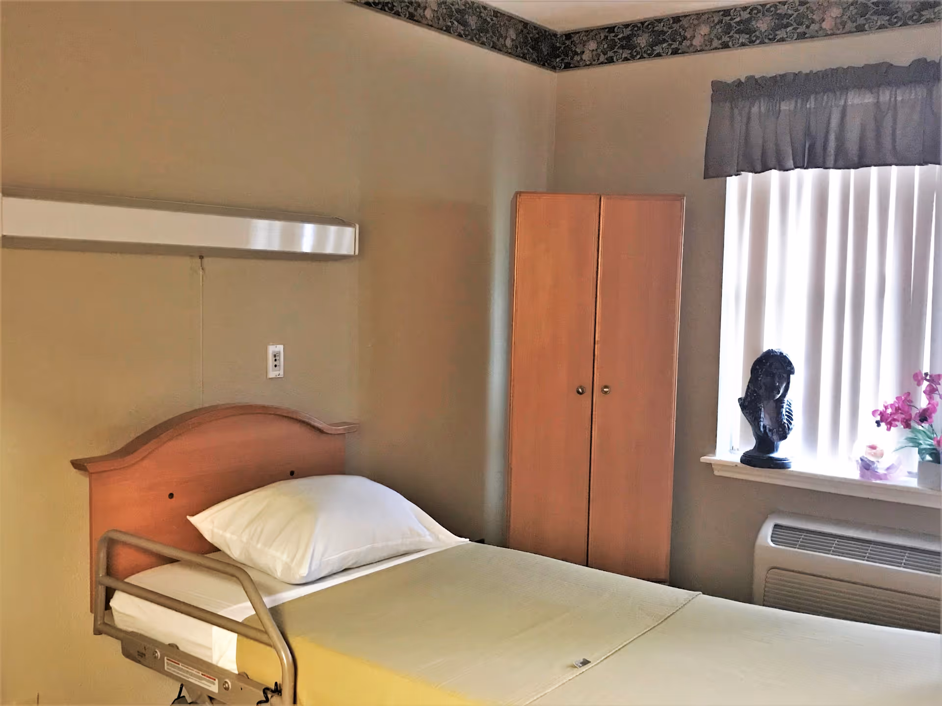 A simple senior living bedroom with a single bed, wooden wardrobe, window with vertical blinds, and a decorative bust on the windowsill.
