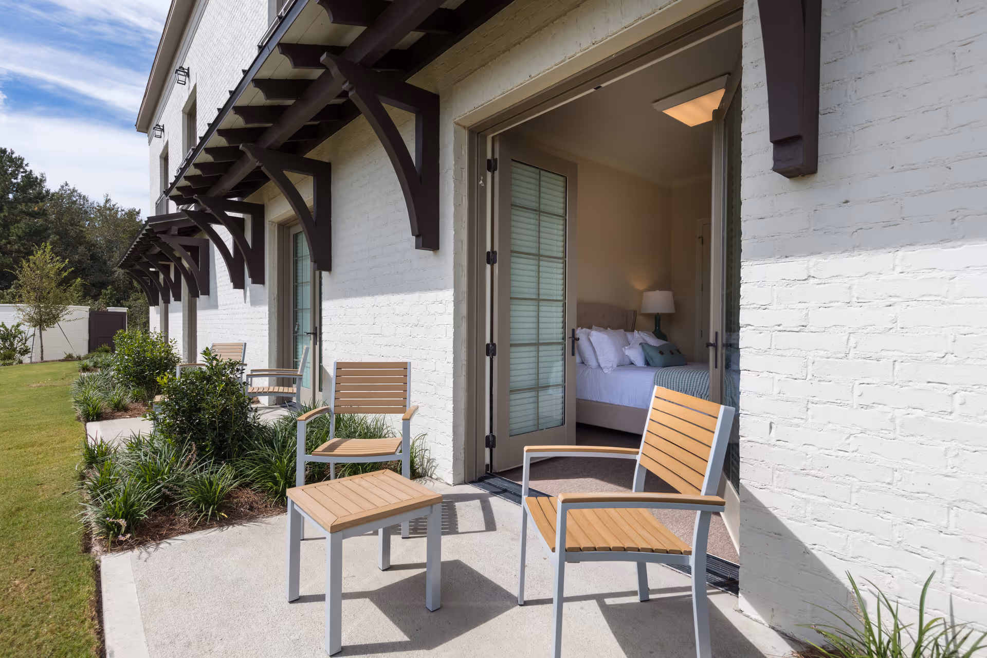 Outdoor patio area with wooden chairs and a small table on a concrete walkway next to a white brick building. The building has large glass doors that open into a bedroom with a bed, pillows, and a bedside lamp. There are plants and grass along the side of the building under wooden awnings.