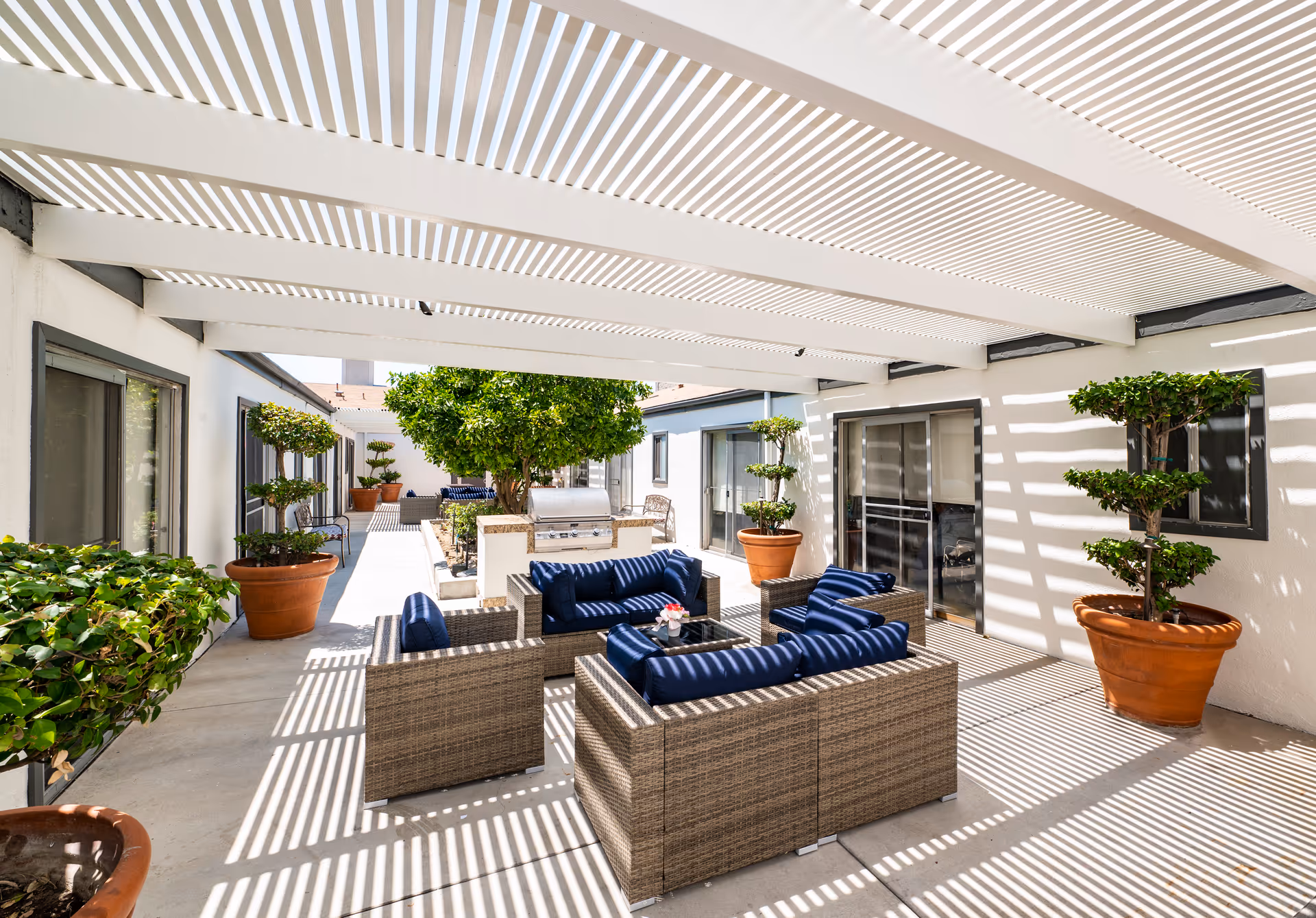 Shaded outdoor courtyard with wicker lounge seating with blue cushions, potted topiary trees, and a pergola casting striped shadows.