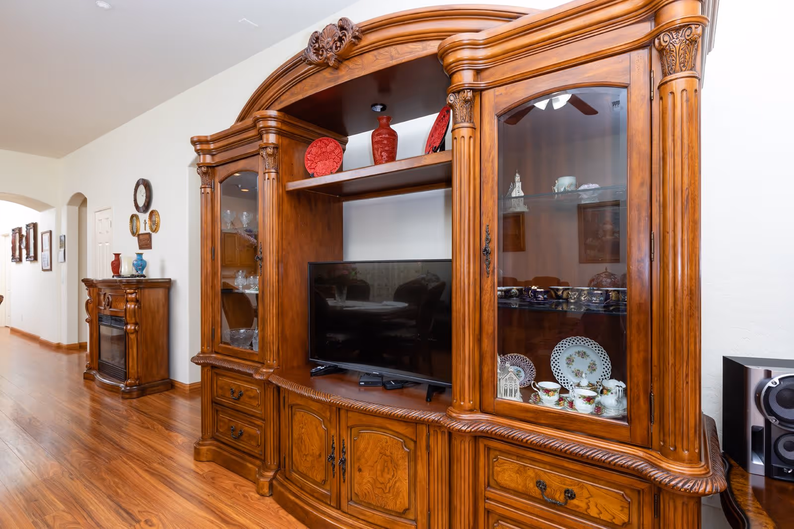 Ornate wooden entertainment cabinet in a living room displaying a TV, decorative plates and vases with hardwood floors and a hallway visible to the left.