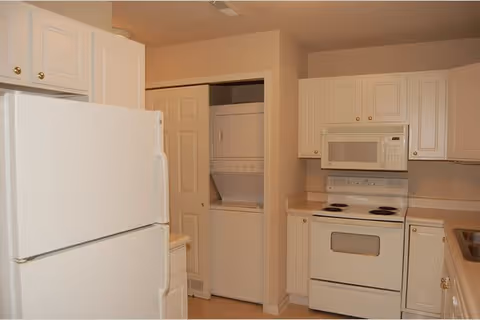 A kitchen with white cabinets, a white refrigerator, a white electric stove with oven, a white microwave above the stove, and a stacked washer and dryer unit in an open closet.