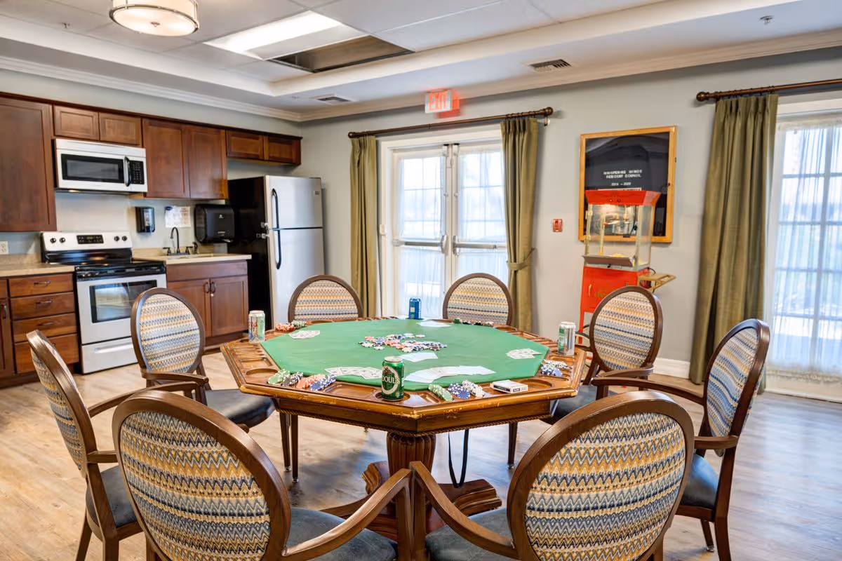 A spacious room with a kitchen area featuring wooden cabinets, a microwave, stove, and refrigerator. In the center, there is a large octagonal poker table with playing cards, poker chips, and soda cans on it, surrounded by eight cushioned chairs with patterned upholstery. The room has large windows with green curtains and a popcorn machine in the corner.