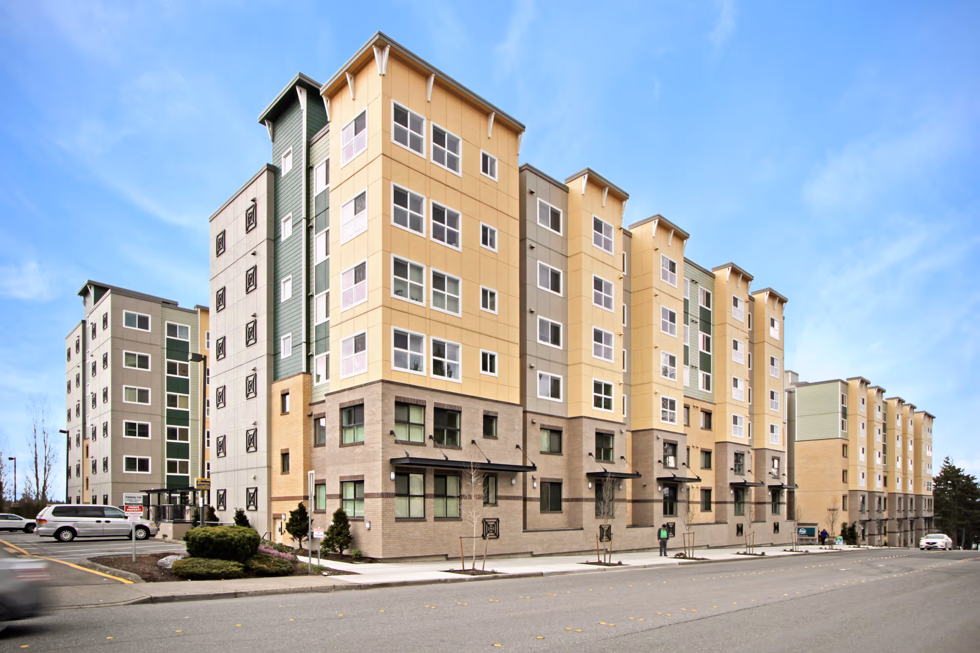 Exterior view of a multi-story residential building with beige, gray, and green panels under a clear blue sky. There are a few small trees and shrubs along the sidewalk, and a couple of vehicles parked nearby.