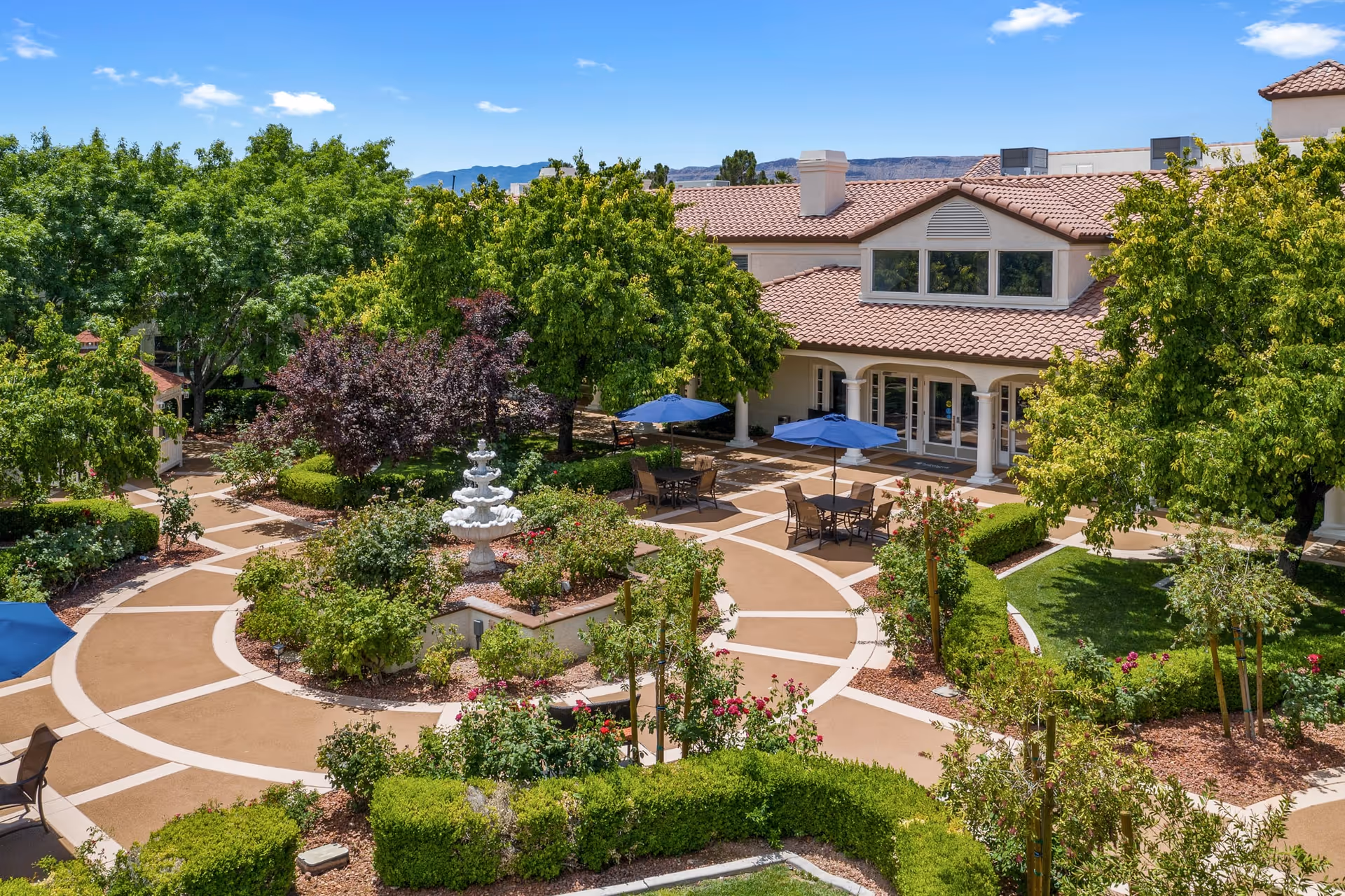 A landscaped outdoor courtyard with a central multi-tiered white fountain surrounded by green bushes, trees, and flower beds. There are several tables with blue umbrellas and chairs arranged on paved walkways. In the background, there is a building with a red-tiled roof and large windows under a clear blue sky.