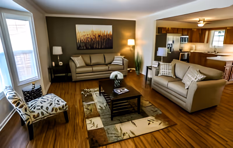 A cozy living room with two beige sofas, a patterned armchair, a wooden coffee table on a decorative rug, a floor lamp, and a painting on the wall. The room has wooden flooring and an open view into a kitchen area with wooden cabinets and stainless steel appliances.