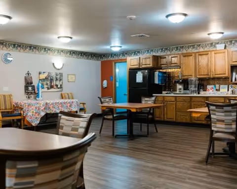 Communal dining area with round tables and chairs, a kitchenette with wooden cabinets and a coffee station, and a table covered with a floral tablecloth.