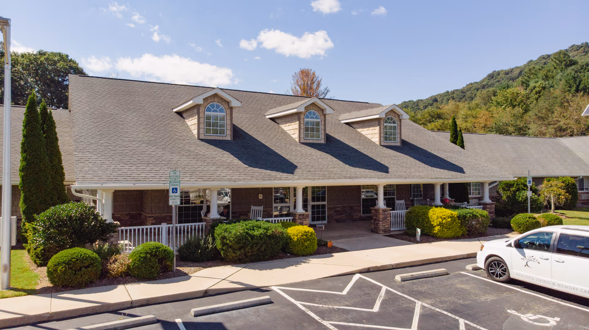 Exterior view of Yancey House senior living facility showing a single-story building with a gray shingled roof, three dormer windows, a covered porch with white railings and rocking chairs, surrounded by neatly trimmed bushes and trees under a partly cloudy sky.