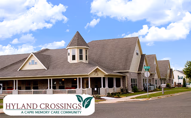 Exterior view of a large residential-style building with a steep roof and a turret, under a blue sky with scattered clouds. The building has a covered porch with white columns and is located at the corner of Liberty Avenue and another street. There is a sign in the foreground that reads 'Hyland Crossings A Capri Memory Care Community.'