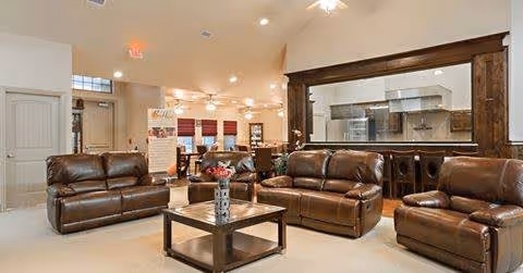A spacious living room area with brown leather sofas and a matching armchair arranged around a wooden coffee table with a small decorative item on it. The room has beige walls and carpet, recessed lighting, and a large window-like opening with wooden trim that looks into a kitchen area with stainless steel appliances.