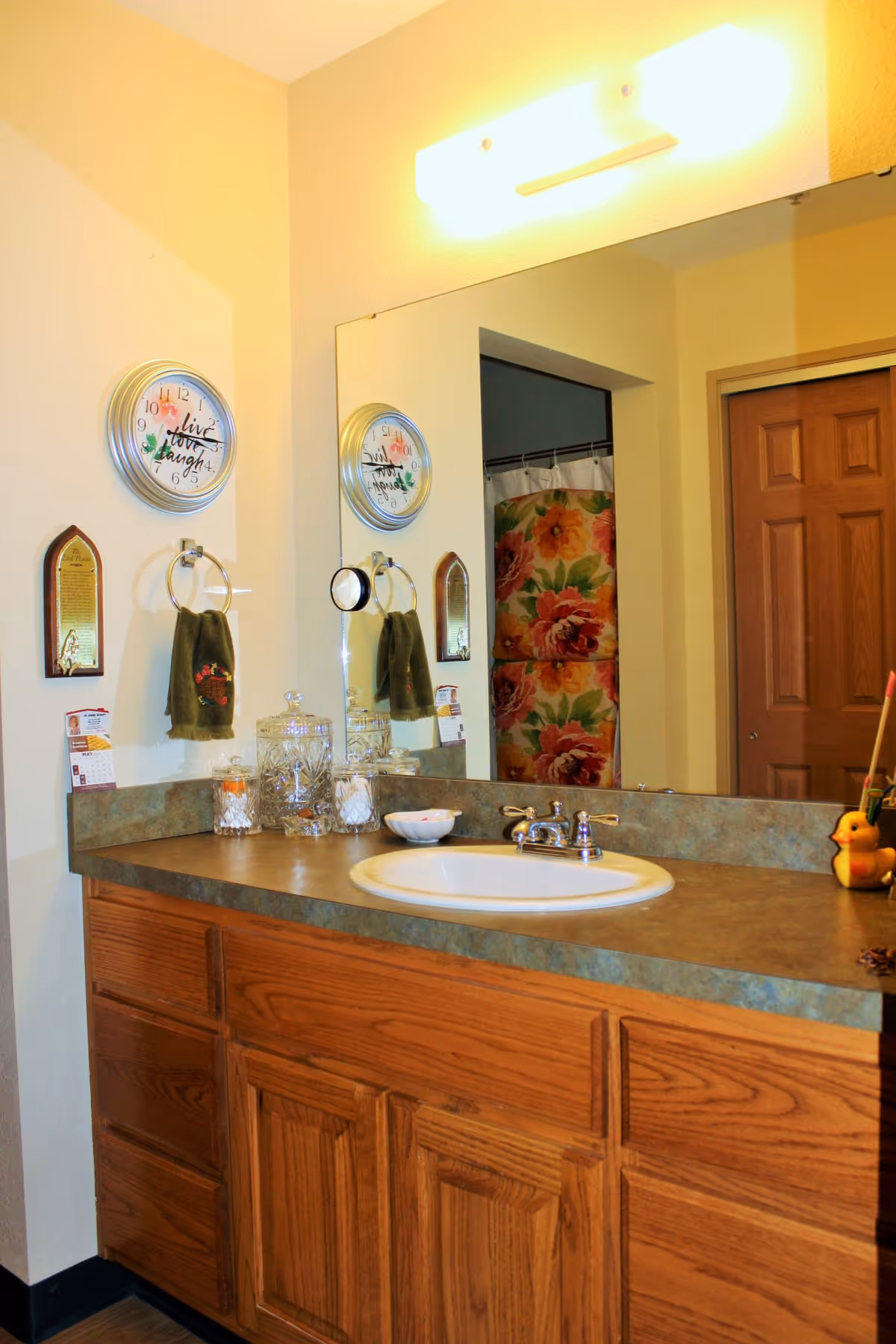 Bathroom vanity with a wooden cabinet, a white sink, and a large mirror above. On the countertop are glass jars, a small bowl, and a yellow rubber duck with a toothbrush holder. A wall clock with the words 'Live, Love, Laugh' hangs on the wall next to a towel ring holding a green towel. In the reflection of the mirror, a floral shower curtain and a wooden door are visible.