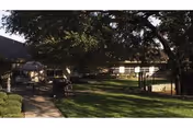 Outdoor view of a senior living facility courtyard with a large tree providing shade, a paved walkway, benches, and a fenced area. The building is visible in the background with windows and a sloped roof.