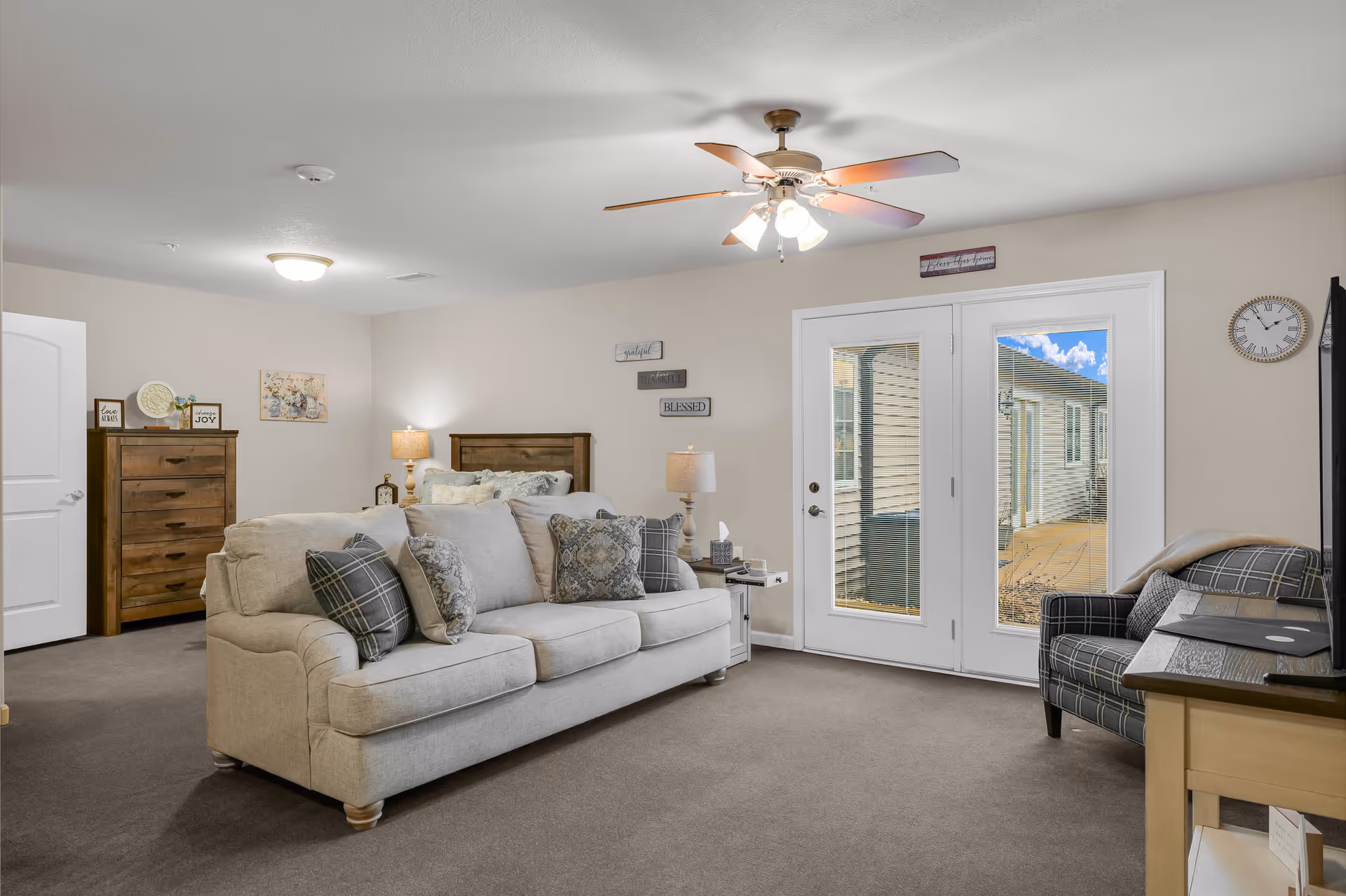 Bright living room with a beige sofa and patterned armchair, ceiling fan, dresser and French doors leading outside.