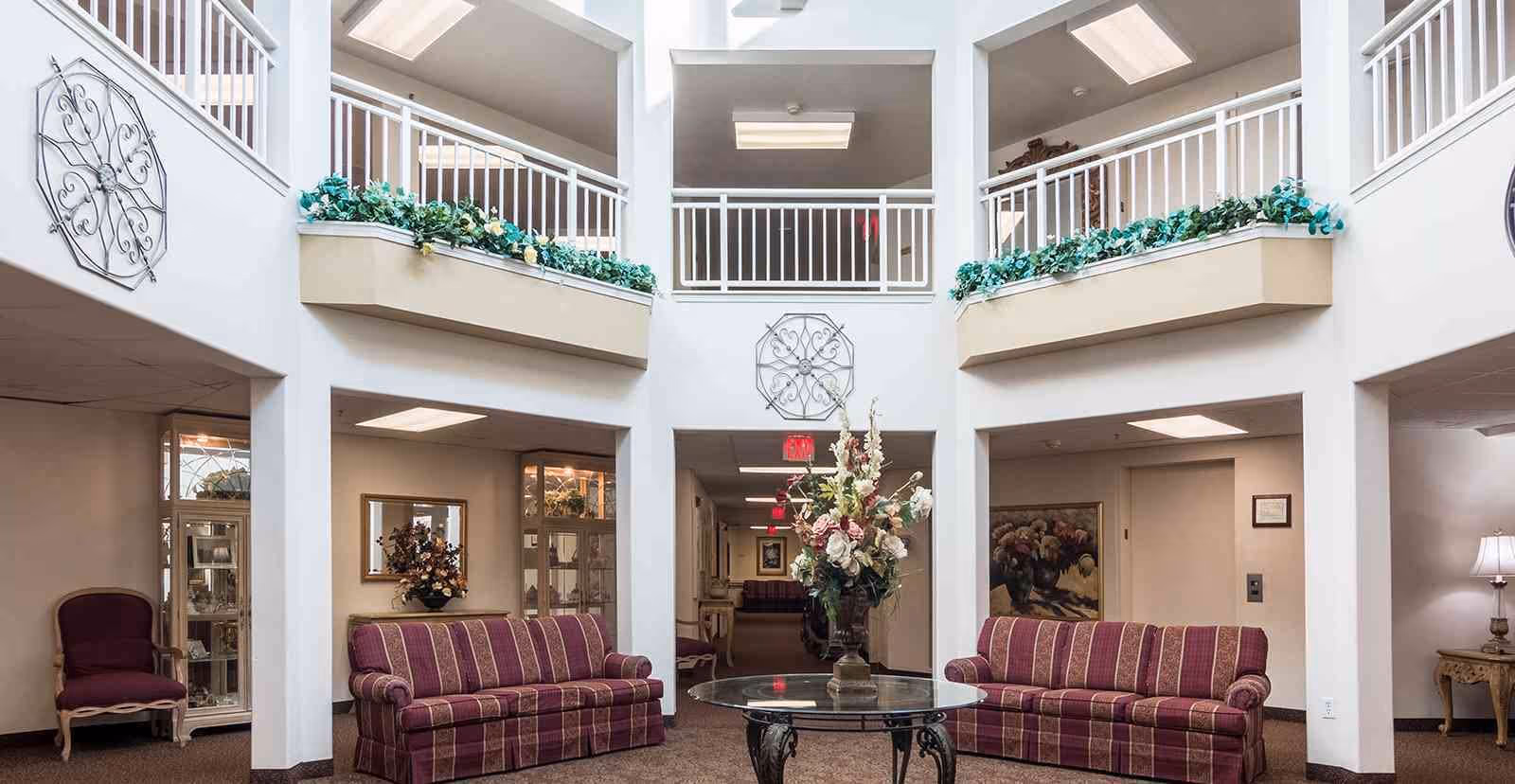 A bright and spacious common area in a senior living facility featuring two striped purple sofas facing each other with a round glass table holding a large floral arrangement in the center. The room has white walls, decorative metal wall art, and an upper balcony with white railings and planters filled with greenery. There are additional seating areas and lamps visible in adjacent rooms.