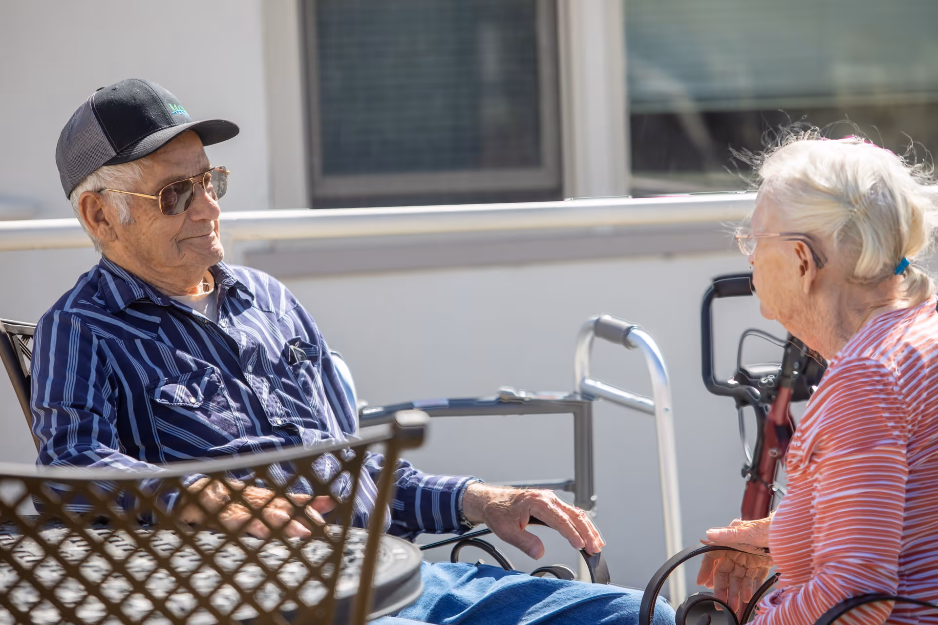 An elderly man wearing a striped shirt, sunglasses, and a cap is sitting outdoors and conversing with an elderly woman in a striped shirt. Both are seated near a metal table, and mobility aids such as a walker and a rollator are visible nearby.