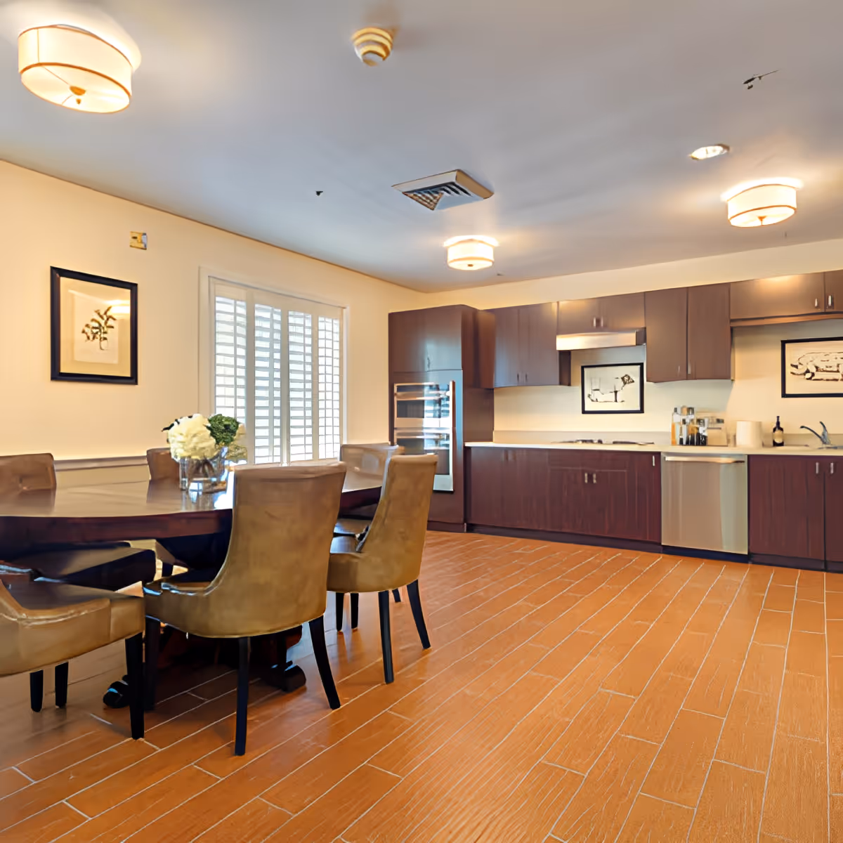 A spacious kitchen and dining area with a round wooden dining table surrounded by six brown leather chairs. The kitchen features dark wood cabinets, stainless steel appliances including a double oven and dishwasher, and framed artwork on the walls. The room is well-lit with ceiling lights and natural light coming through white plantation shutters on the window.