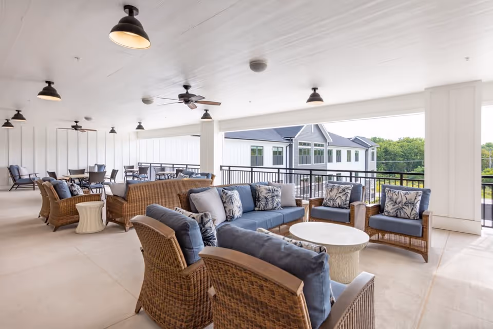 Covered outdoor patio area with wicker seating including sofas and armchairs with blue cushions and patterned pillows, round white tables, ceiling fans, and hanging lights. The patio overlooks a building and greenery in the background.