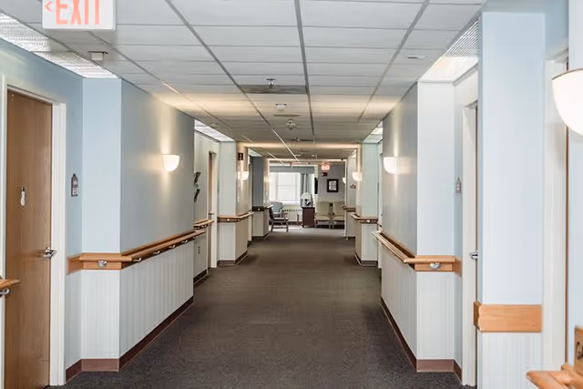 A long, well-lit hallway in a senior living facility with light blue walls, wooden handrails on both sides, multiple closed doors, and a seating area with chairs and a table visible at the far end.