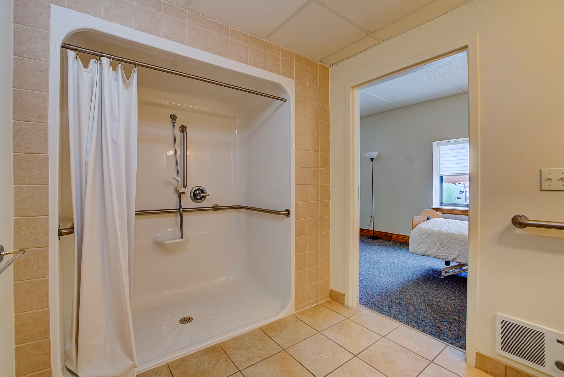 Accessible tiled bathroom with a walk-in shower and grab bars, looking into an adjacent bedroom.