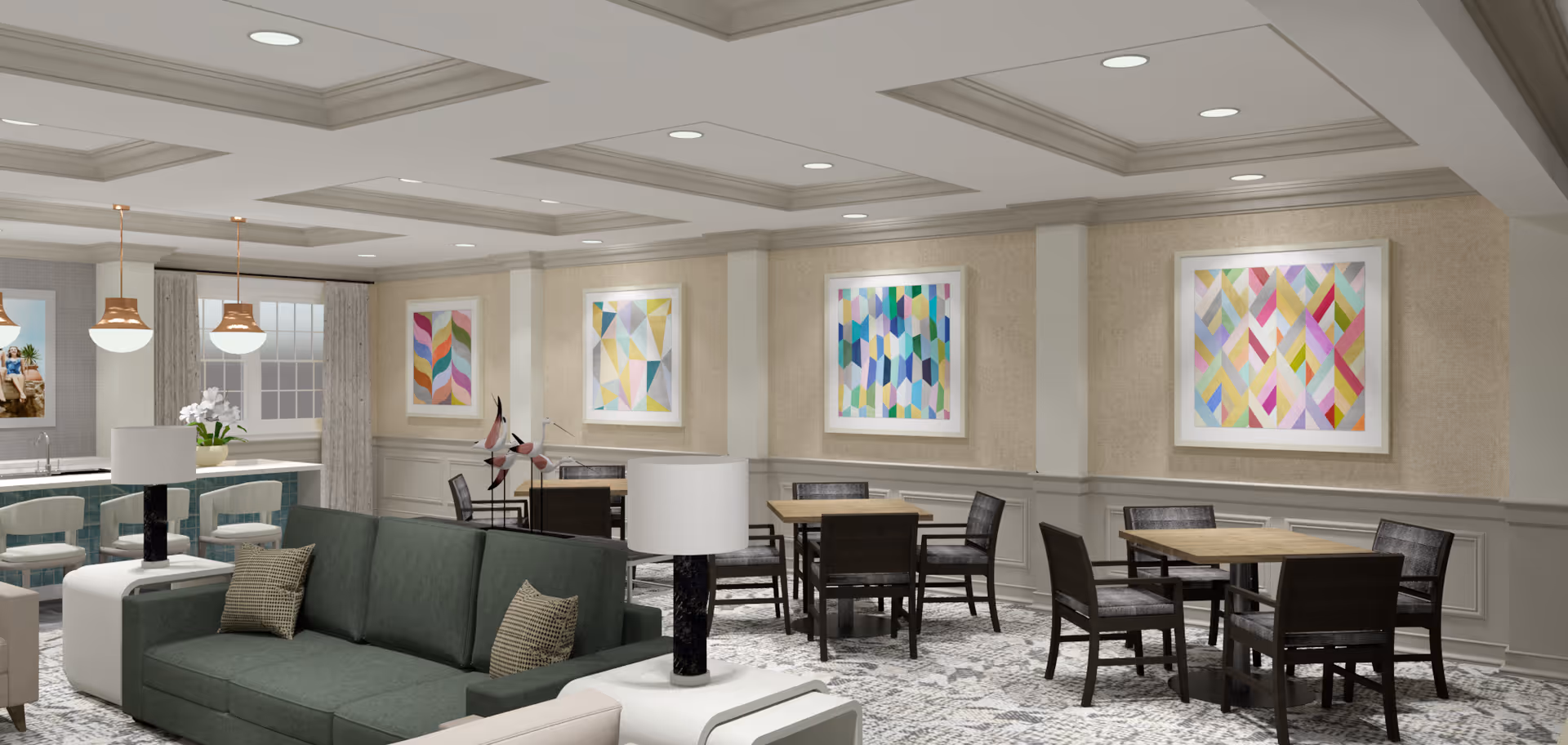 A spacious and well-lit common area with a green sofa, white side tables with lamps, and several wooden tables with chairs. The walls are decorated with colorful abstract paintings, and there are pendant lights hanging over a counter area in the background.