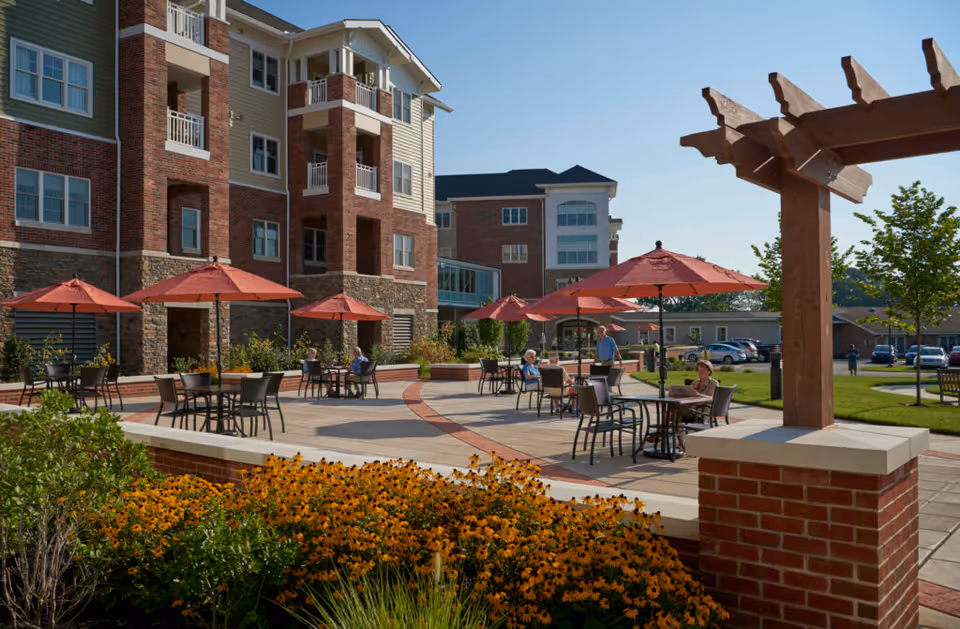 Outdoor patio area at Landis Homes with several tables and chairs under red umbrellas. People are sitting and standing around the tables. The patio is surrounded by a brick and stone building, with flower beds and green grass nearby under a clear blue sky.