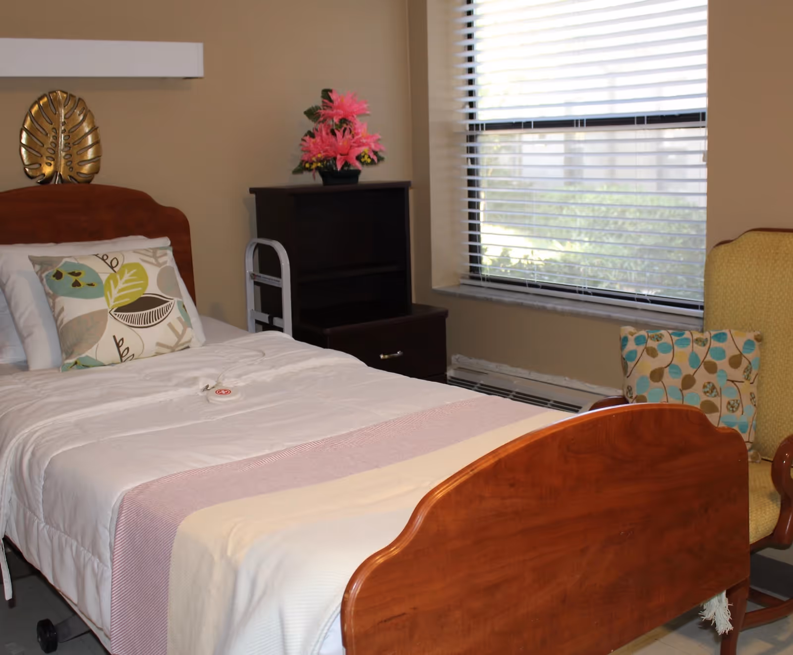 A neatly made single bed with white and light pink bedding, a decorative pillow with leaf patterns, and a wooden headboard and footboard. Next to the bed is a dark wooden nightstand with a pink flower arrangement on top. A window with white blinds is on the right side, and a yellow armchair with a patterned cushion is partially visible beside the window.