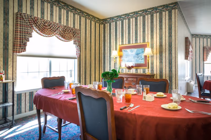 Dining room with a long table covered with a red tablecloth, set with plates, glasses of iced tea, and silverware. The room has striped wallpaper with floral patterns, a window with plaid valances, a wooden sideboard with two lamps and a framed picture above it, and carpeted floor.