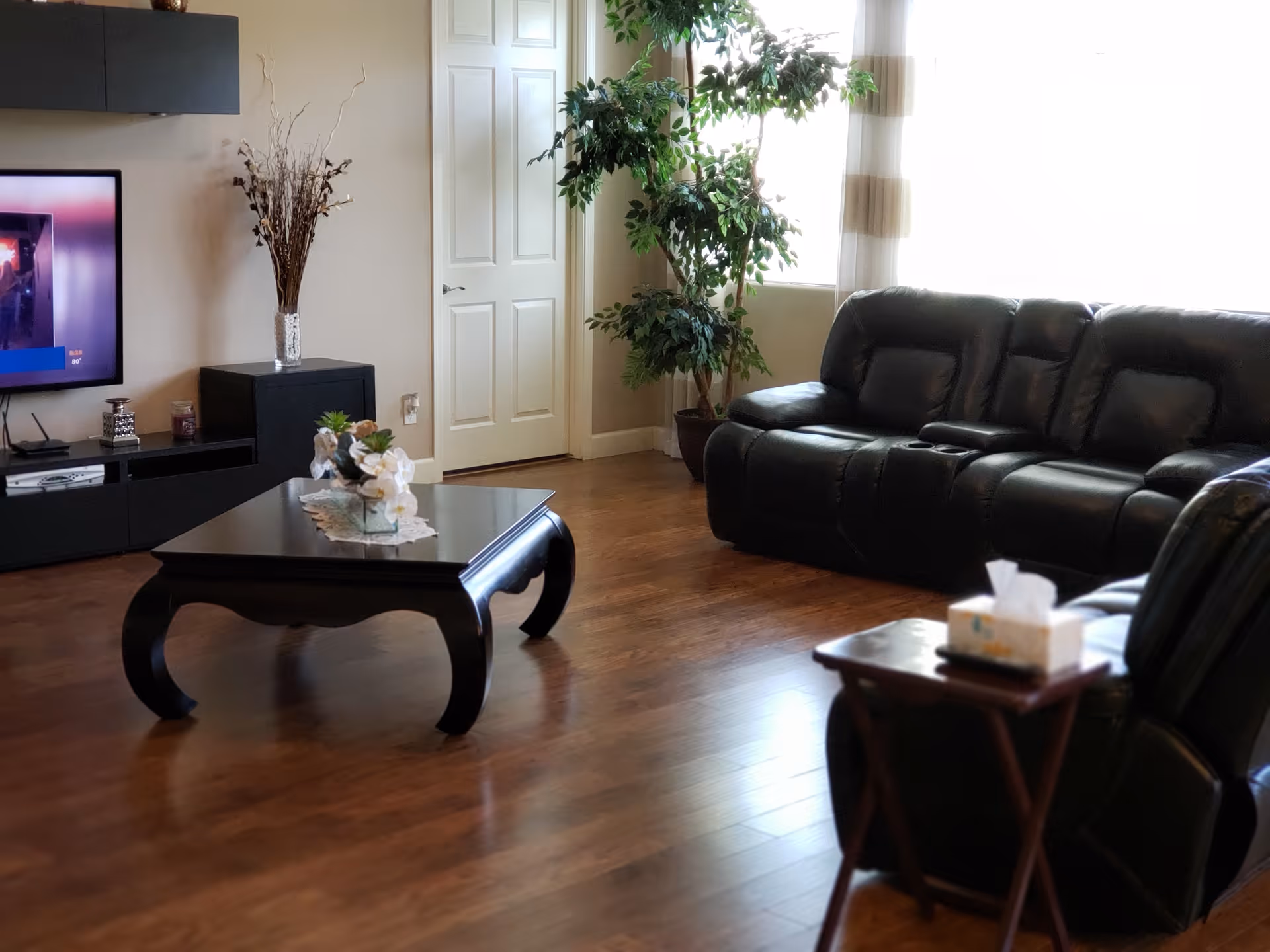 Spacious living room with black leather sofas, a dark wooden coffee table, TV stand, and a potted plant on hardwood floors.
