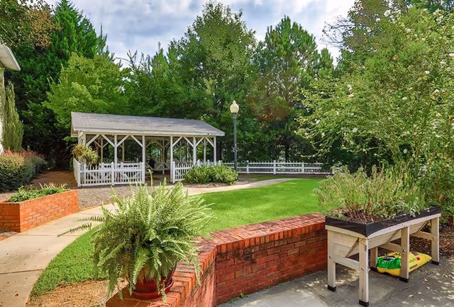 Outdoor garden area with a covered pavilion, green lawn, brick planter walls, a potted fern, and a raised garden bed with plants. Trees and shrubs surround the space under a partly cloudy sky.