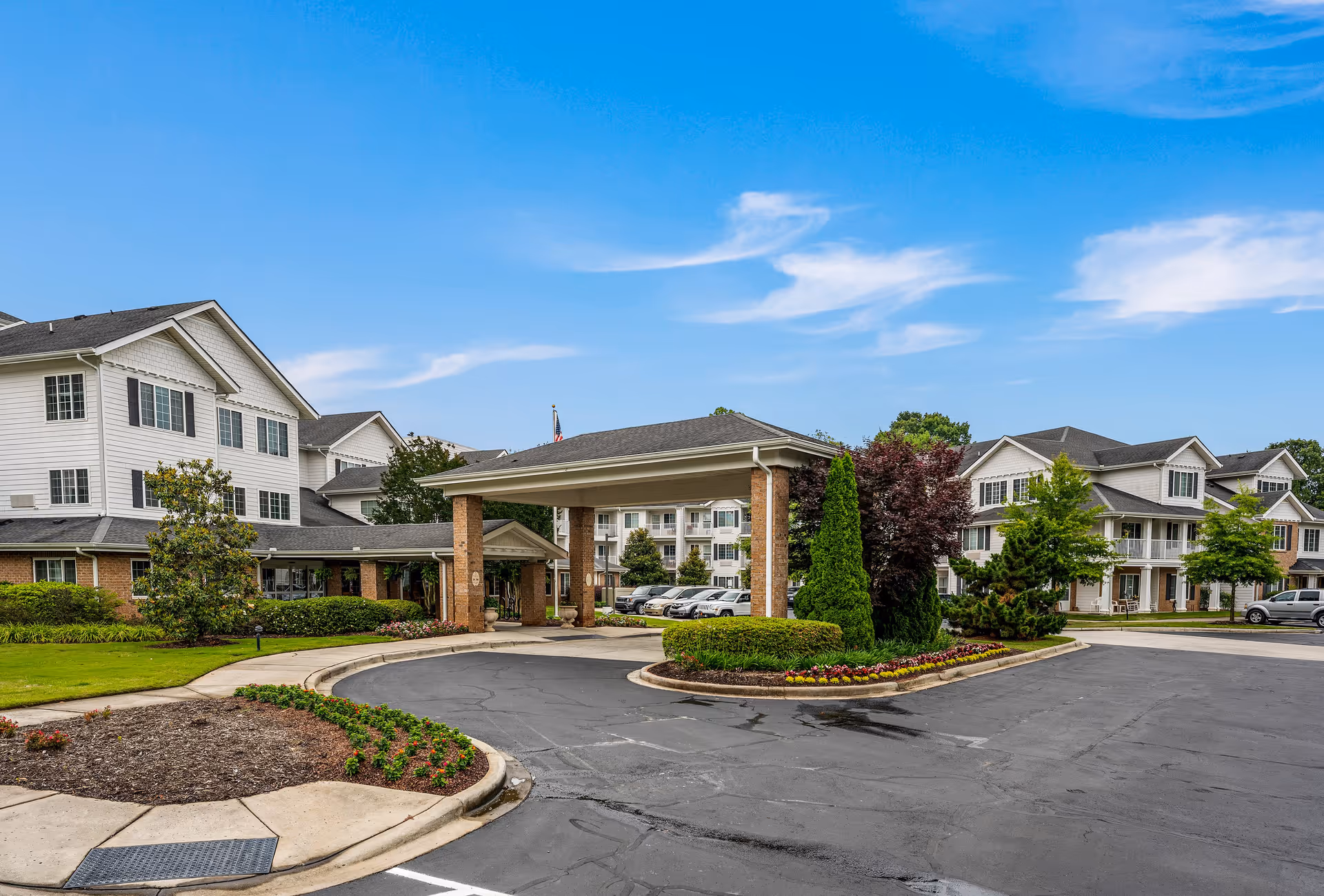 Exterior view of Shads Landing Gracious Retirement Living facility showing a covered entrance with brick pillars, landscaped greenery, and multiple residential buildings under a blue sky.