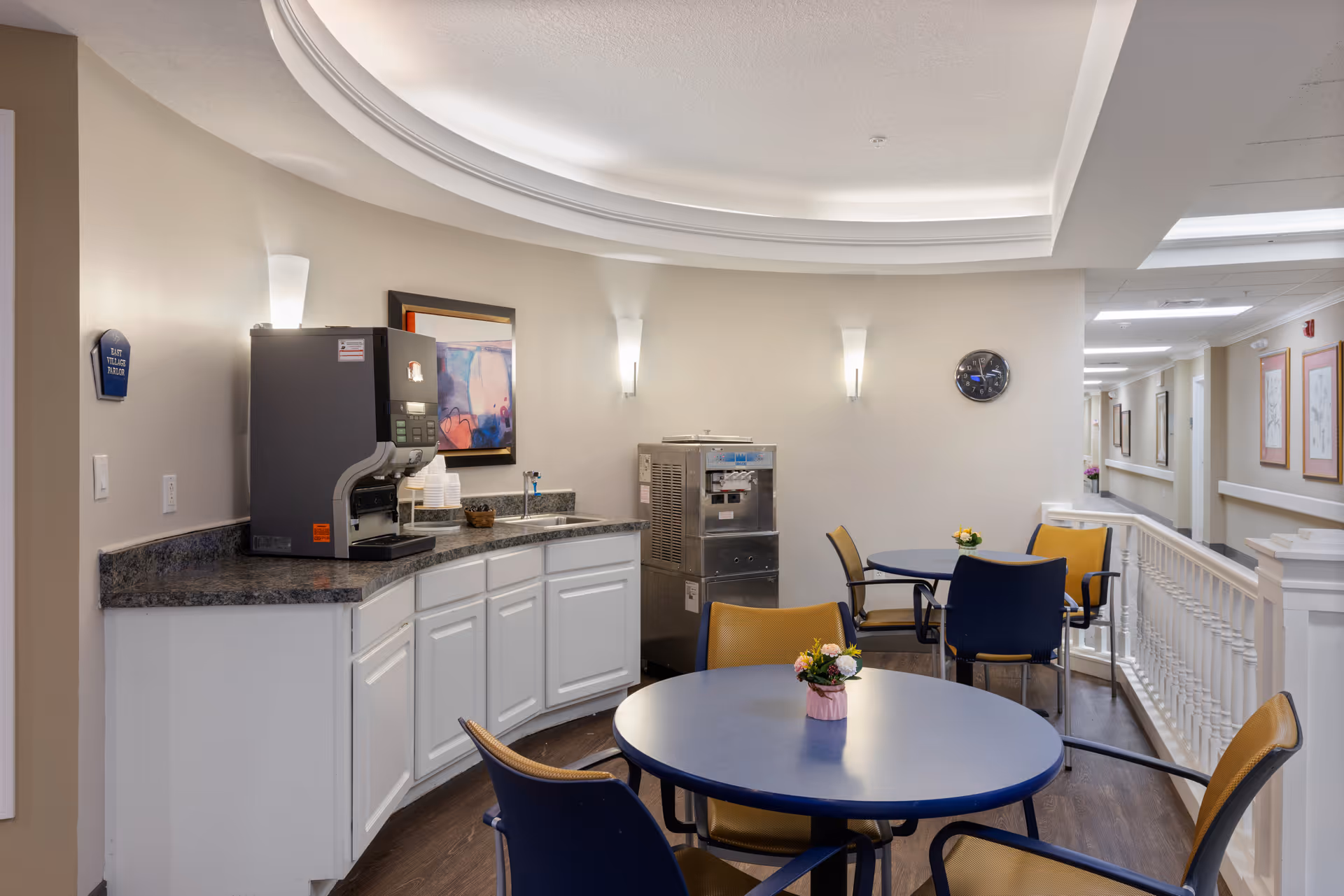 A small seating area in a senior living facility with round tables and chairs. There is a countertop with a coffee machine and an ice cream or frozen yogurt dispenser. The walls are light-colored with modern wall sconces and a framed abstract painting. A clock is mounted on the wall, and a hallway with framed artwork is visible in the background.