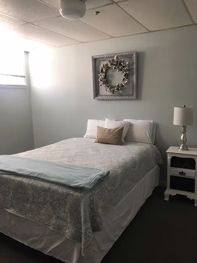 A neatly made bed with a patterned bedspread and a small decorative pillow with the word 'love' on it in a simple bedroom. There is a white nightstand with a silver lamp on the right side of the bed. A framed wreath decoration hangs on the wall above the bed. A window with blinds is partially visible on the left side.