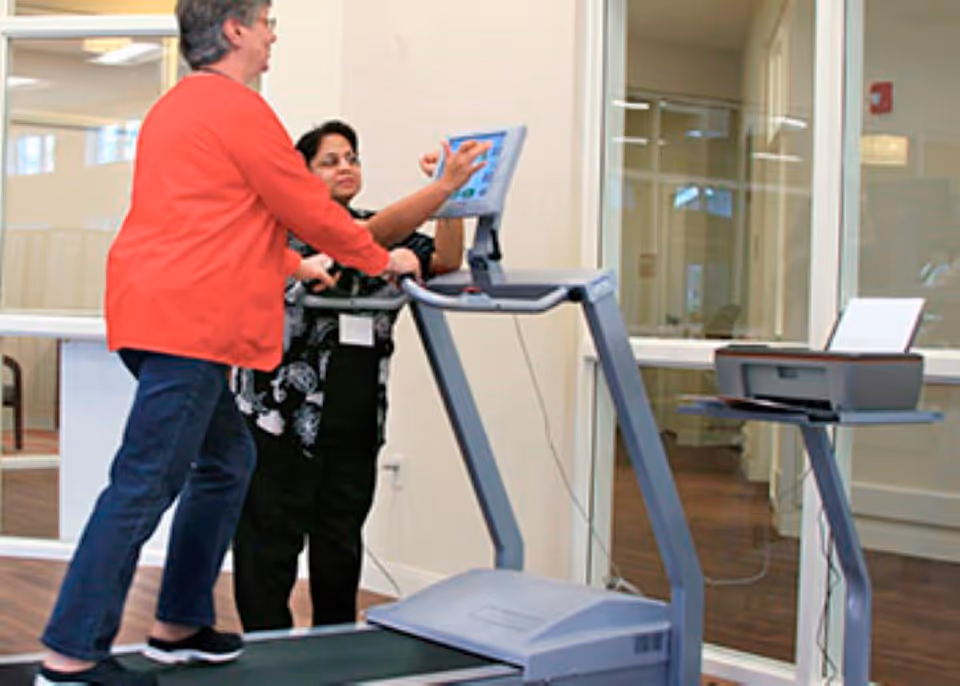 An elderly woman in a red jacket is walking on a treadmill while a staff member in a black outfit assists her by pointing at the treadmill's control panel in a well-lit indoor setting with wooden floors and large windows.