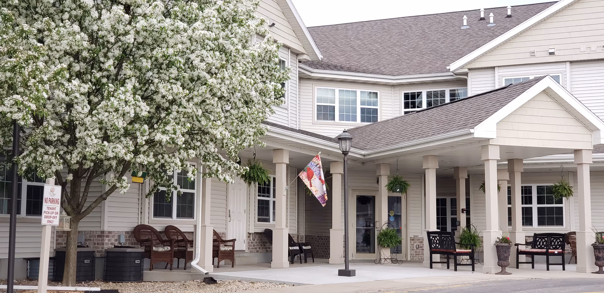 Exterior view of The Meadows Of Fall River, L.L.C. facility showing a covered entrance with columns, outdoor seating with benches and chairs, hanging plants, a blooming tree with white flowers, and a no parking sign for tenant pick-up or drop-off only.