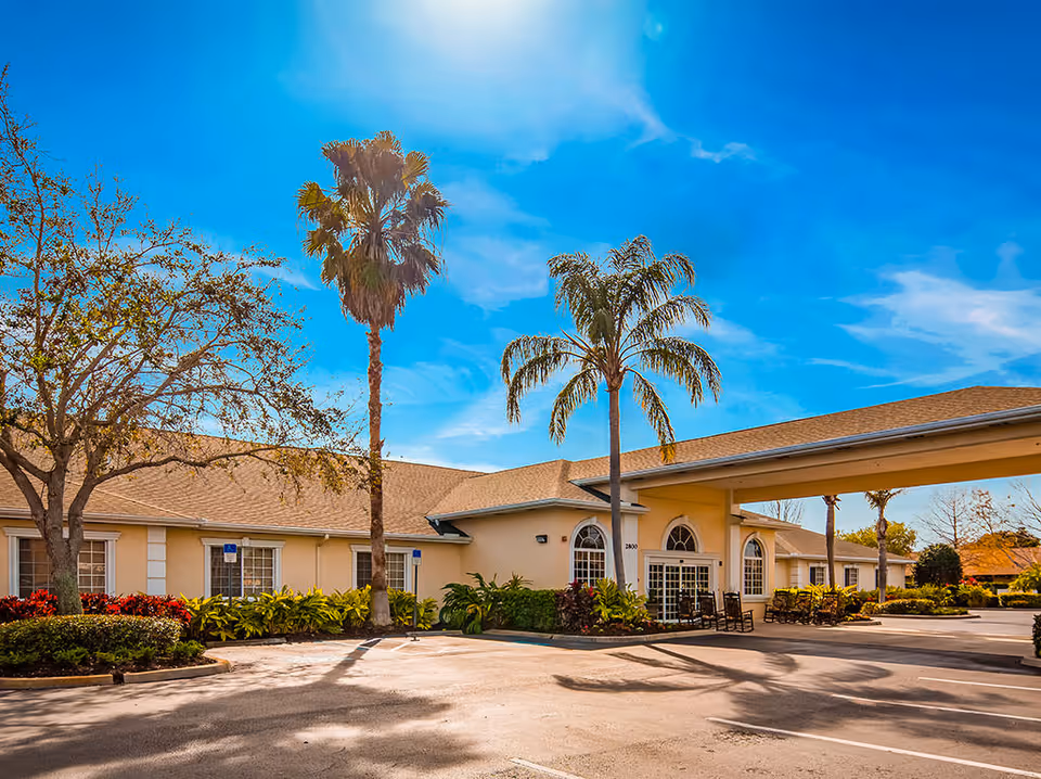 Exterior view of The Windsor of Bradenton senior living facility on a sunny day, showing a single-story building with a covered entrance, palm trees, and landscaped greenery around the parking area.