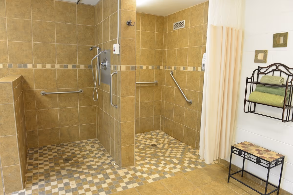 A spacious, accessible shower area with two open shower stalls featuring beige and brown tiled walls and floors. Each stall has a grab bar and a handheld showerhead. To the right, there is a metal rack mounted on the wall holding folded green towels, and a small matching bench below it. A beige shower curtain is partially visible.