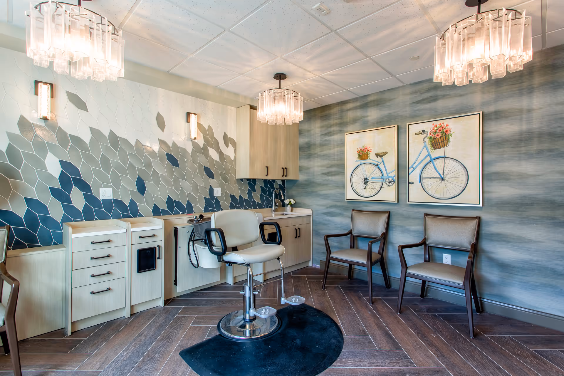 A modern salon area with a styling chair in the center, two guest chairs against a blue textured wall with two framed bicycle paintings, light wood cabinetry with a sink, and a decorative hexagonal tile backsplash in shades of blue and gray. The room has two elegant ceiling light fixtures and wood-patterned flooring.