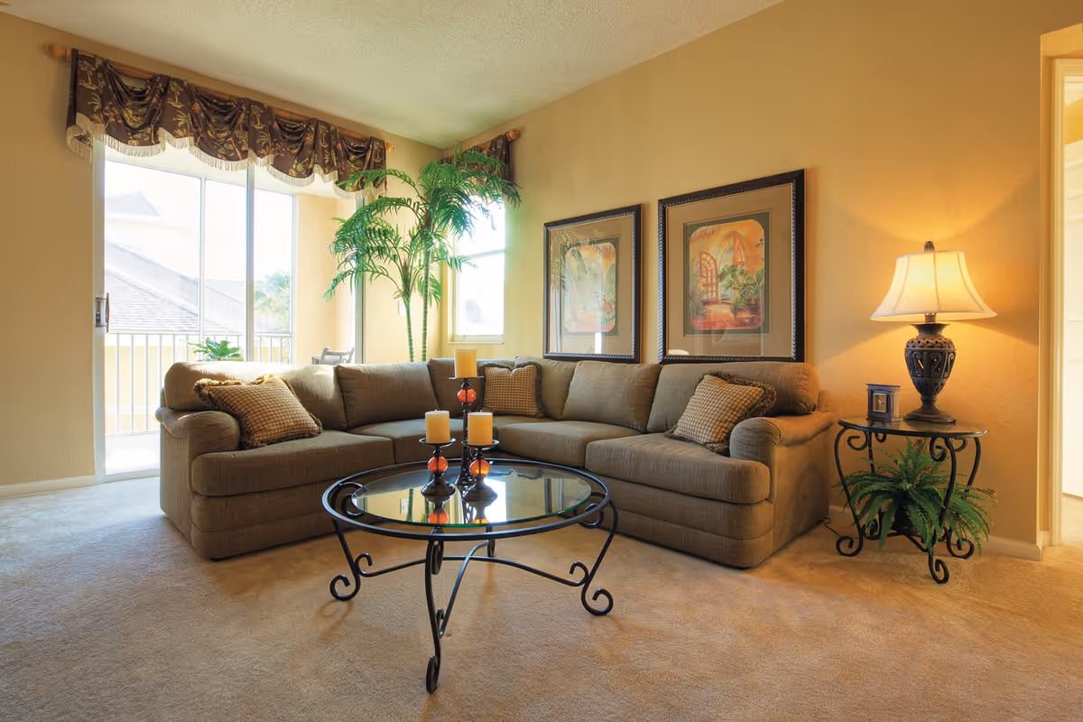 A cozy living room with a large beige sectional sofa adorned with patterned cushions, a glass coffee table with three candles, two framed paintings on the wall, a side table with a decorative lamp and a small plant, and a tall potted plant near a sliding glass door with a floral valance.