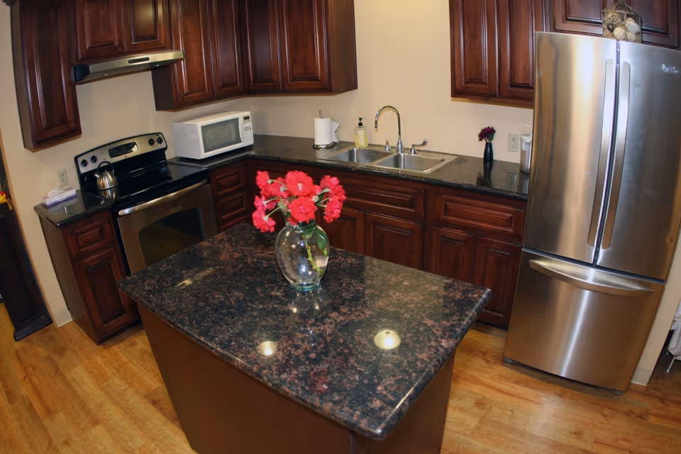 A kitchen with dark wooden cabinets, a stainless steel refrigerator, stove, and microwave. There is a double sink with a faucet, a paper towel holder, and a soap dispenser on the countertop. In the center, there is a kitchen island with a dark granite countertop and a glass vase holding red flowers. The floor is wooden.