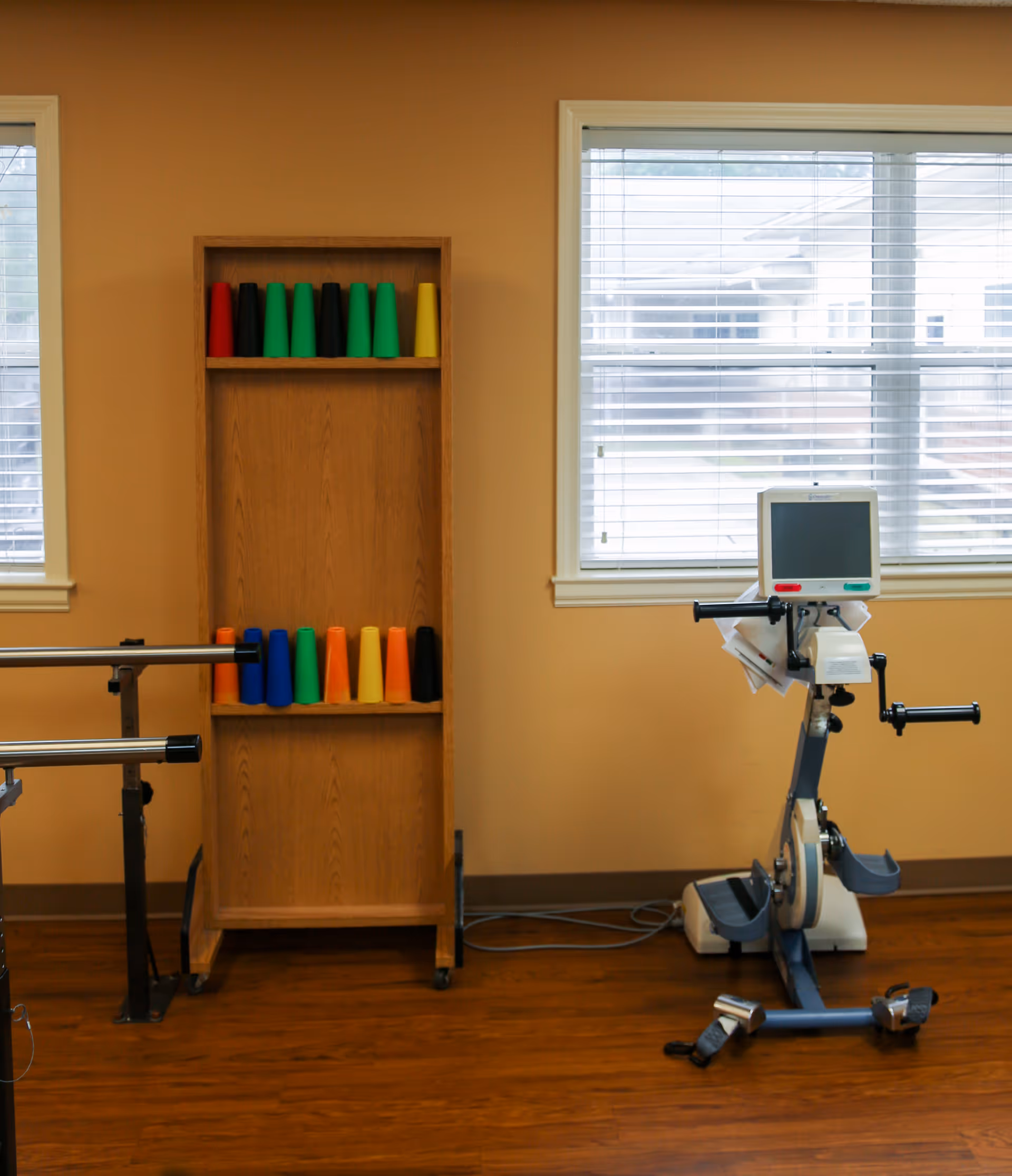 Physical therapy room with parallel bars for walking support, a wooden shelf holding colorful cones, and a stationary exercise bike with a digital monitor in front of two windows with blinds.