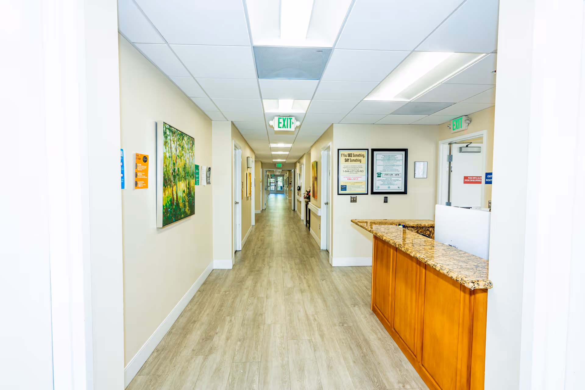 Long, well-lit interior hallway of a senior living facility with wood-look floors, artwork on the walls and a reception desk on the right.