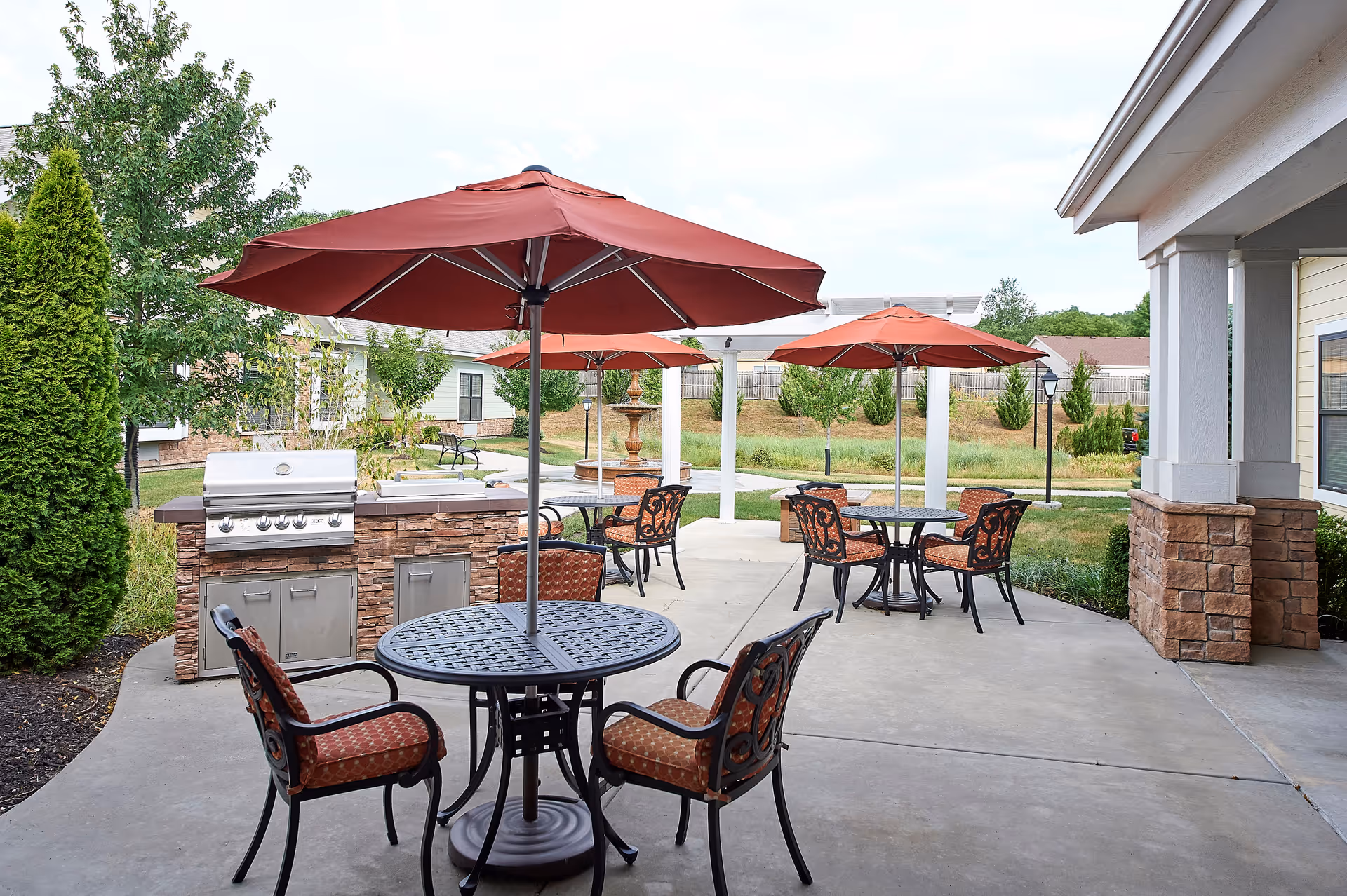Outdoor patio area with round metal tables and cushioned chairs under large red umbrellas. There is a built-in stone grill and countertop area, surrounded by greenery and trees, with a fountain and pergola in the background.